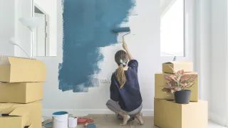 Woman painting wall blue; boxes and plant in room.