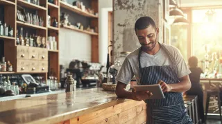 Barista smiling, using a tablet, leaning on a bar.