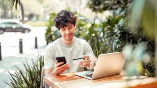 Younger man sitting at table with computer and phone, entering numbers off of a card into phone