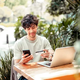 Younger man sitting at table with computer and phone, entering numbers off of a card into phone